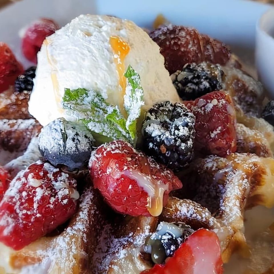 Golden waffle topped with whipped cream, fresh berries, powdered sugar, and syrup, with a side of syrup.