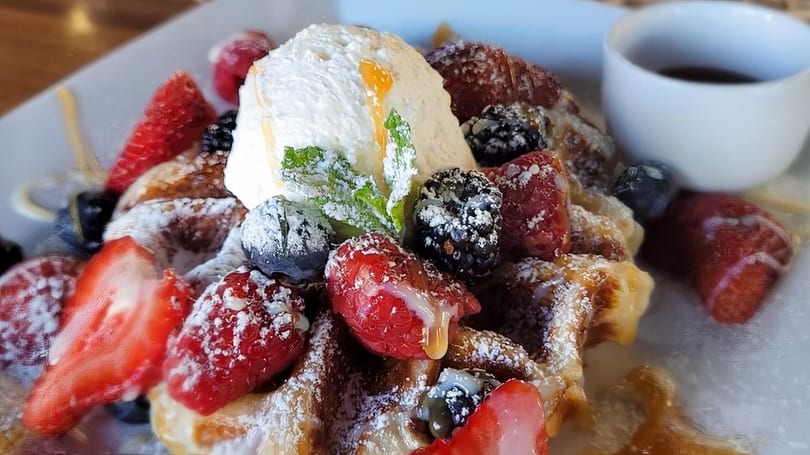 Golden waffle topped with whipped cream, fresh berries, powdered sugar, and syrup, with a side of syrup.